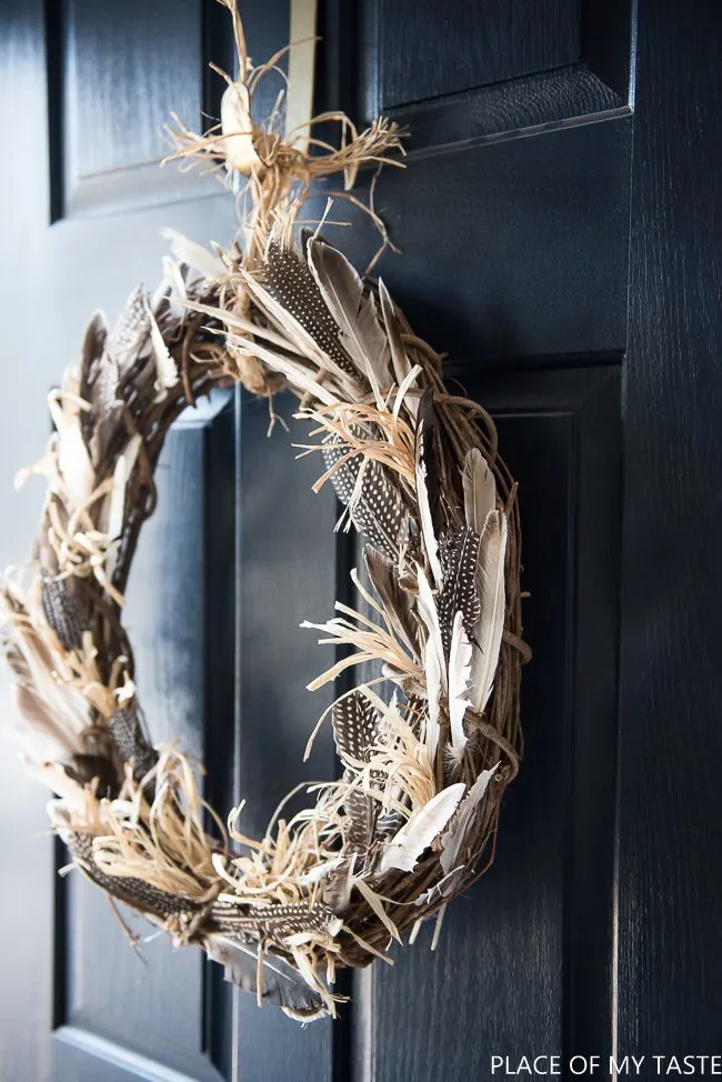 fall wreath ideas made of a grape vine wreath and decorated with raffia and beautiful spotted feathers and gray feathers and hung by raffia or twine on a dark colored black or navy door.