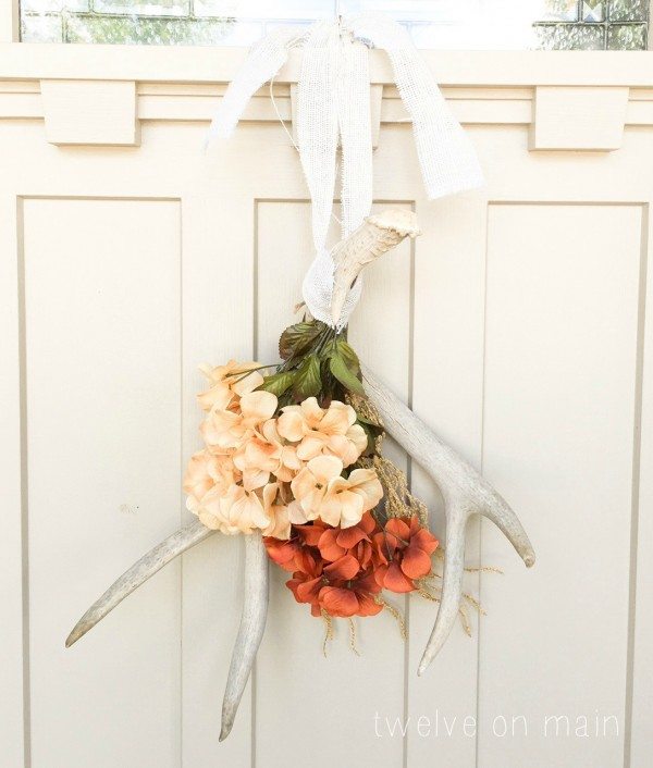 fall wreath ideas antlers hung upside down on a door that is off white.  The antlers have two arrangements of flowers hanging on the front of them. The flowers are off white and a light rust red color.