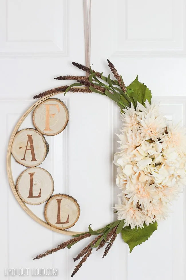 Fall wreaths created on a hoop and on one side are greenery and white flowers and on the inside of the other side is wood slices with gold letters on each wood slice that spells F A L L