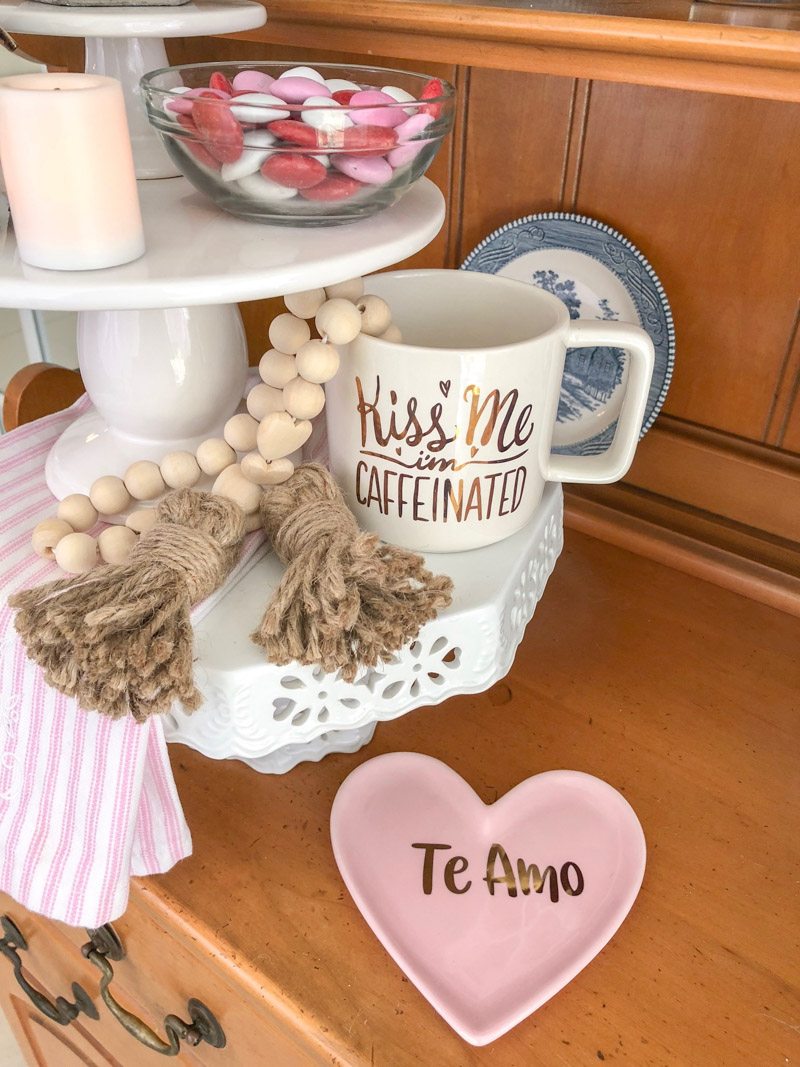 Styling tiered trays with cake stands layered and filled with Valentine decor like a kiss me coffee cup, wooden beads, m&m's in a glass bowl, candles, kitchen towel and more.