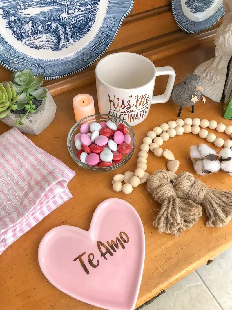 Styling tiered trays with Valentine accessories like a pink and white stripe dish towel, small glass bowl of pink, red and white m&m's, wooden beads, kiss me coffee mug, candle, plant and more.