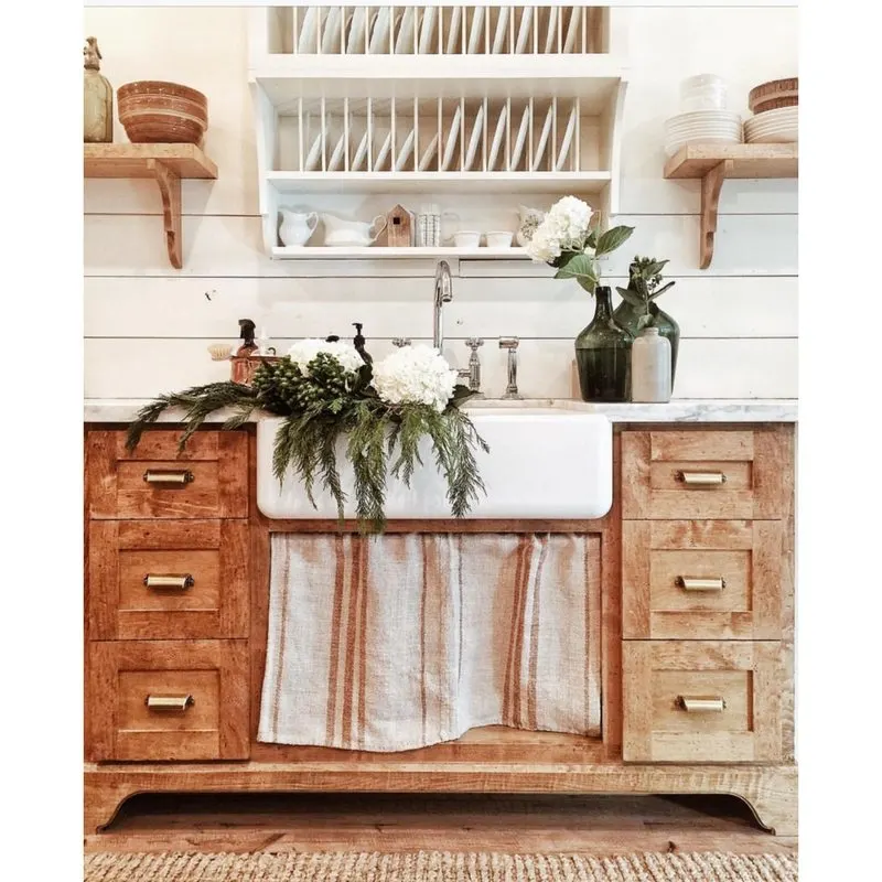 Farmhouse Kitchens by Whitetail Farmhouse with an apron sink and wood cabinetry and flowers in the sink and slatted open shelves with dishes in it above the sink