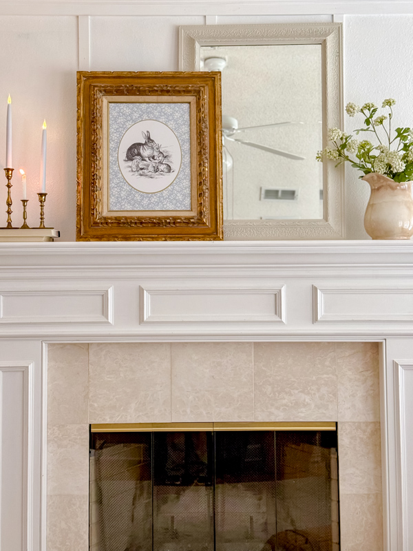 Fireplace mantel decorated for spring using printable art, vintage pitcher with flowers, candles and mirror. The printable art is a cottagecore style blue and white floral mat with a black and white bunny drawing.
