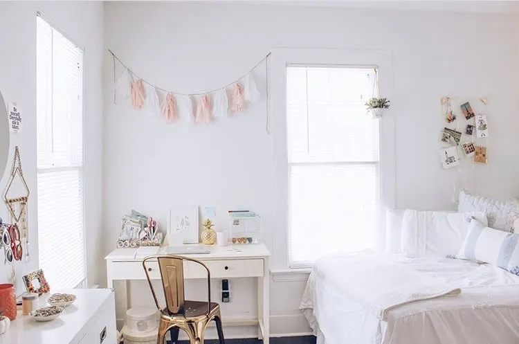 College Dorm Decor Stylin Cafe photograph.  White desk with gold farmhouse style metal chair.  Garland of pink and white paper tassels over desk.  White bedding with white pillows and white throw on bed.  