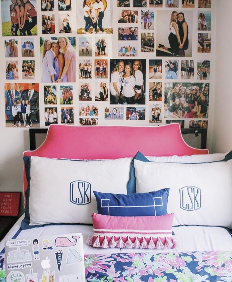 College Dorm Decor Daily Dose of Prep picture.  Wall over bed filled with photographs of friends, Large hot pink pillow that looks like a headboard, which pillow shams trimmed in navy blue and monogrammed, blue geometric pillow and pink accent pillow with tassel fringe.  Apple laptop sitting on the bed filled with stickers like Vineyard Vine and a Lilly Pulitzer throw on the foot of the bed.