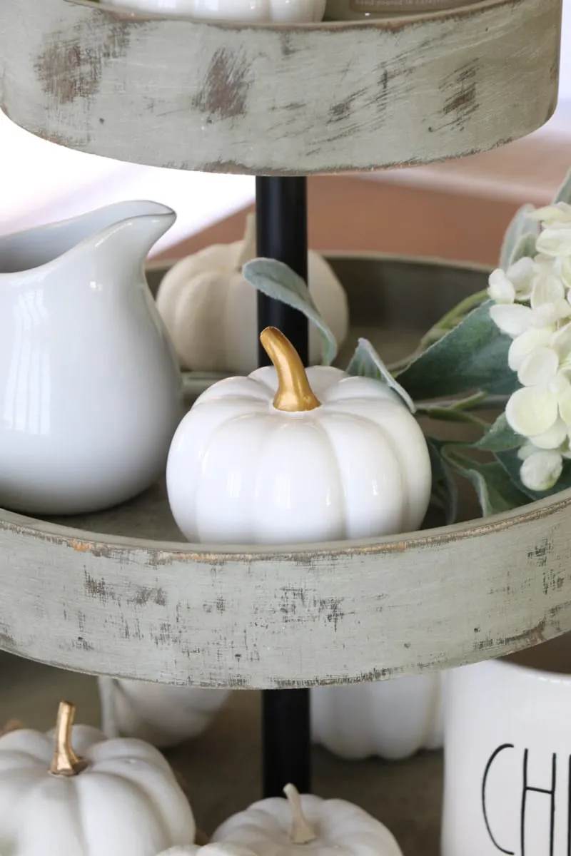 3 tiered wooden stand filled with harvest decorations like white creamer and pumpkin as a centerpiece on a dining table