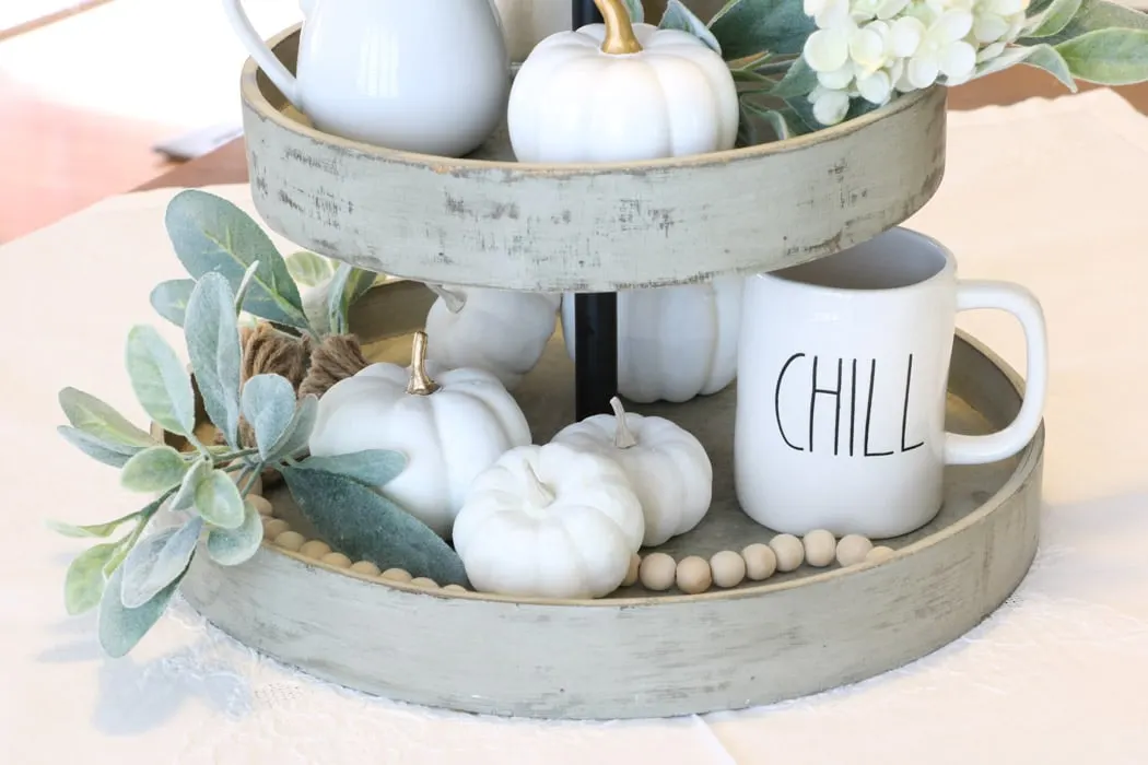 3 tiered wooden stand filled with harvest decorations like white pumpkins, wood bead garland as a centerpiece on a dining table