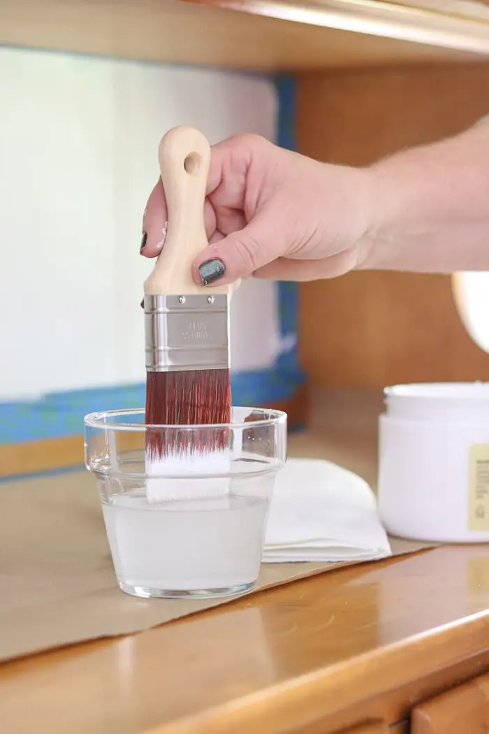 Dipping a paint brush inside a cup of water to keep the brush moist because chalk paint works better if it is moist. 