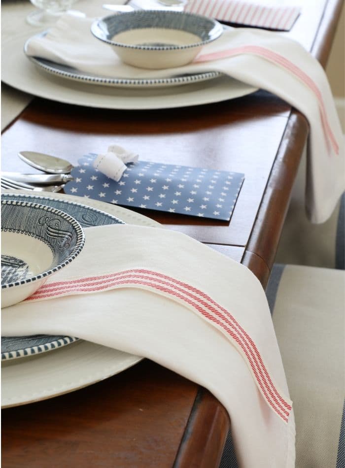 4th of July tablescape with Betsy Ross flags, drop cloth table runner, currier and ives dishes, cloth dinner napkins and red and white ribbon and babies breath 
