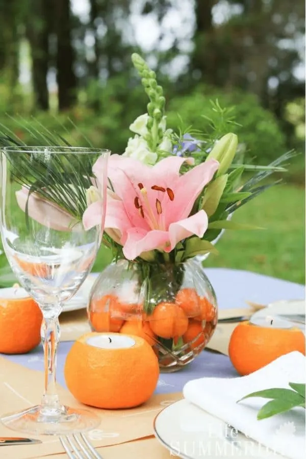 Candle from an orange using a tea light and decorating a table with the orange candles.  this table has a tropical centerpiece with pink followers and greenery placed into a clear vase that also has small oranges in the water.  Around the centerpiece are orange candles with tea lights inside.