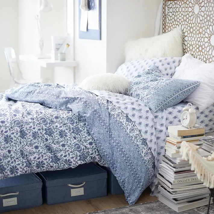 College dorm decor ideas in a pottery barn blue and white paisley comforter set.  Bed is sitting on white painted crates inside a white bedroom.  Ladder on the wall, desk under a window with white paper tassel garland and white industrial lamp.