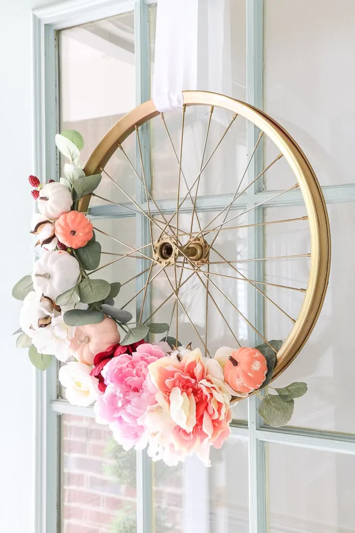Fall decor with a bright colored autumn wreath that incorporates pink on a child's bike wheel that is painted in gold.  