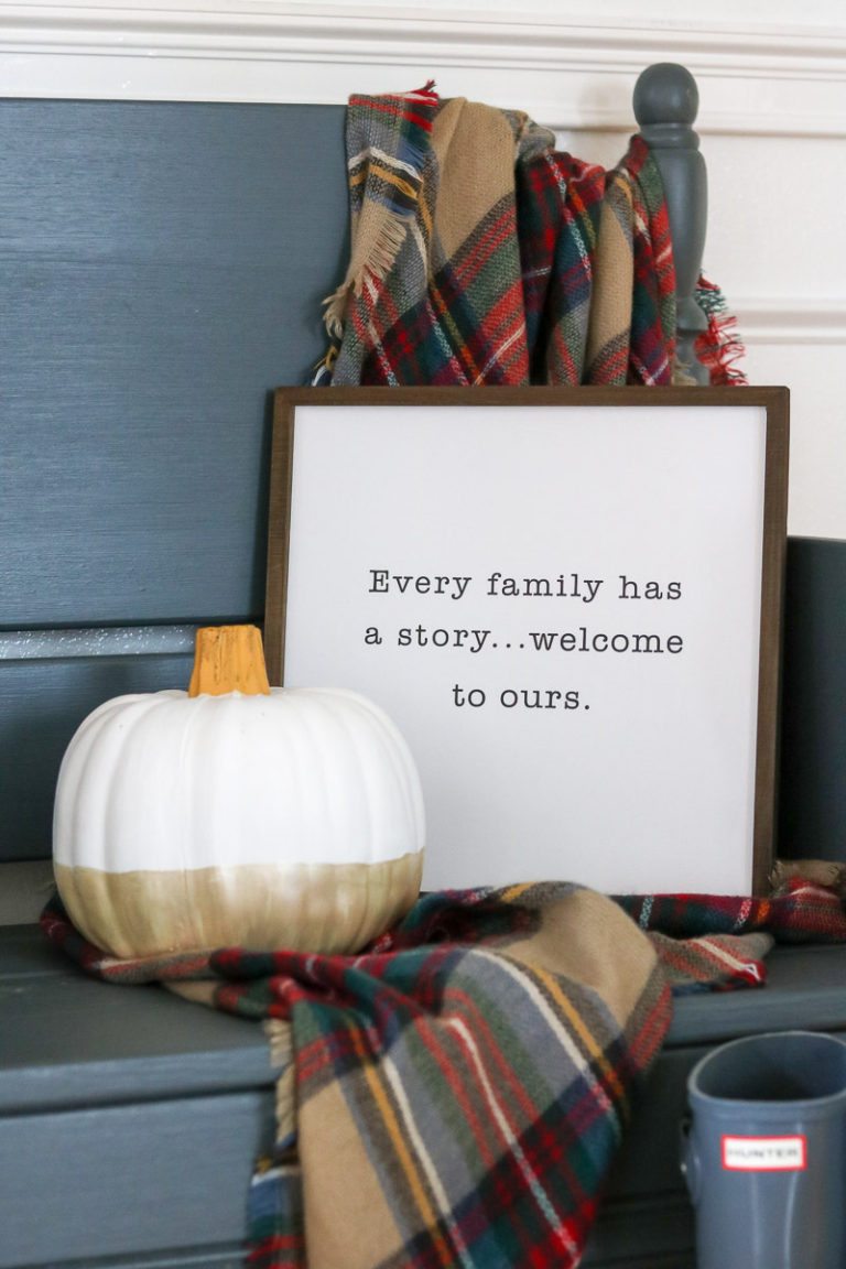 Fall decor ideas using a plaid blanket draped on a blue bench and a sign on the bench that says, "Every family has a story...welcome to ours."  A pumpkin painted champagne gold on the bottom and dusty blue Hunter boots on the floor.