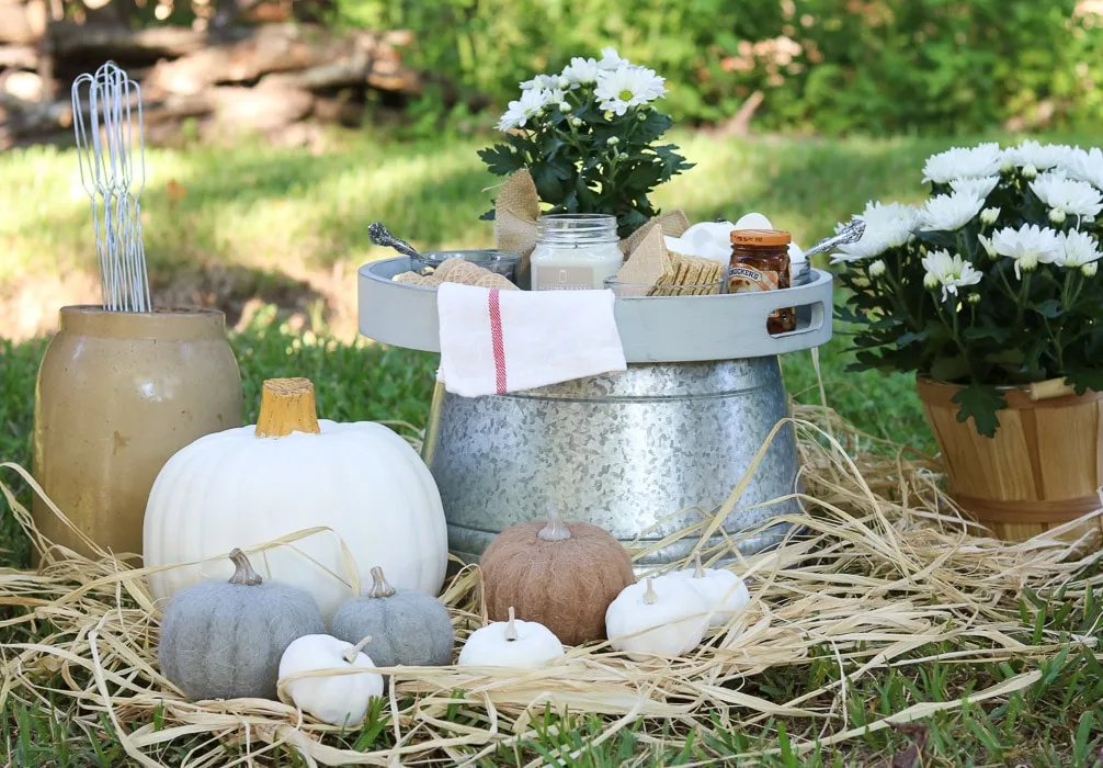 Fall decor idea for the outdoors.  Set up by a fire pit using white mums, pumpkins of different sizes, hay on the ground, a galvanized tub turned upside down and a tray on top filled with s'more items to make s'mores.