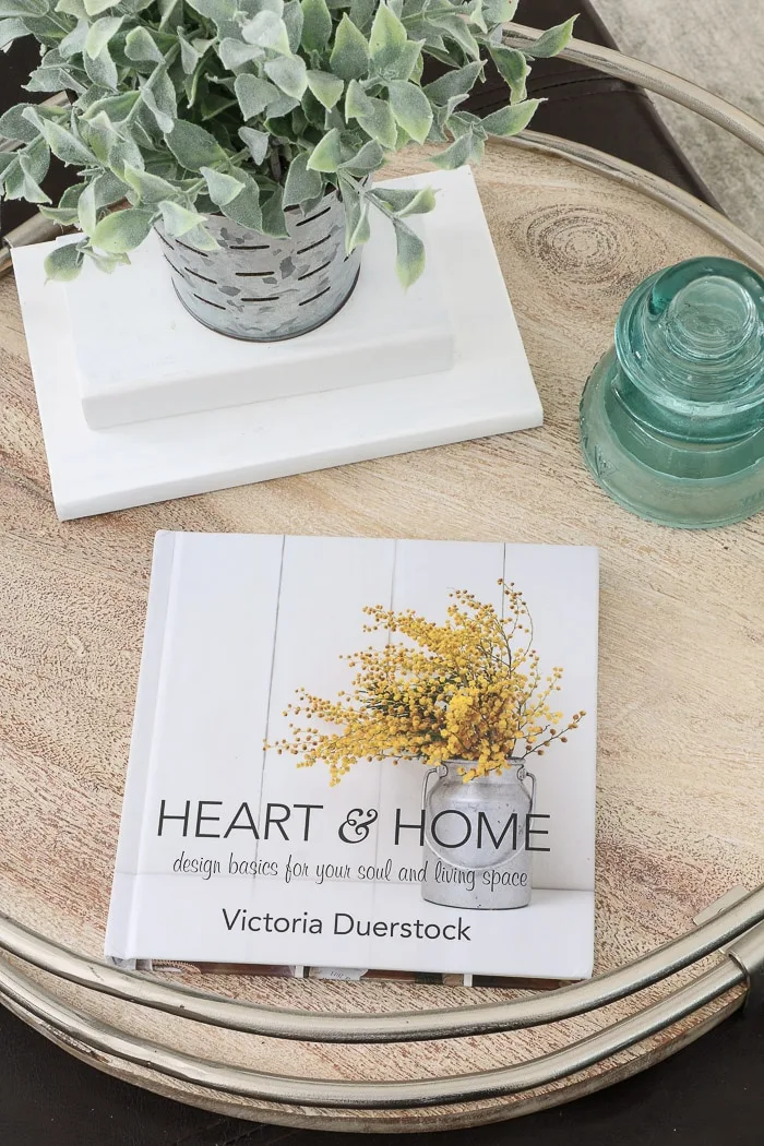 Heart and Home is one of the best interior design books for beginners.  This home decor book and devotional has flowers on the cover and it is sitting with an old insulator, chalk painted books and greenery.