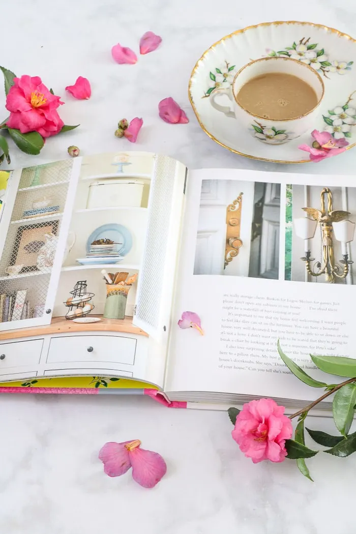 Inside of the book Whiskey in a Teacup by Reese Witherspoon along with a floral china teacup filled with warm coffee and rose colored gardenia flowers and petal sprinkled all around the book.  This is easy one of the best interior design books for beginners.