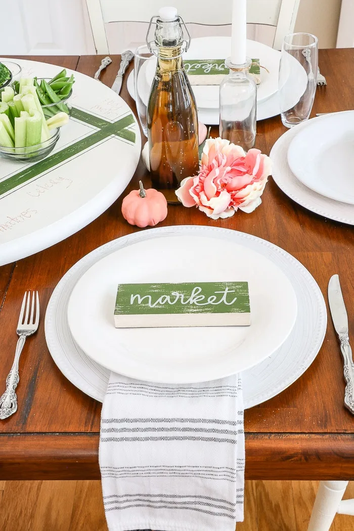 Dollar Tree fall decorating ideas for a Thanksgiving tablescape using a cute market sign for a place setting centerpiece.  Stacked white dishes with a decorative dish towel between the dishes and a white painted charger.