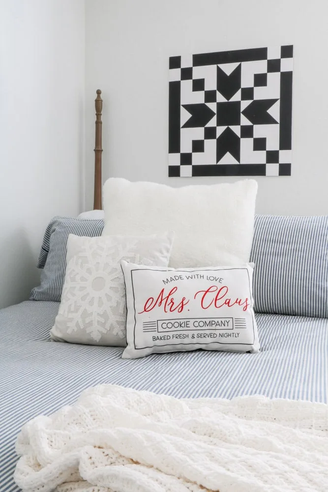 Barn quilt hanging over a bed in a white room with a blue and white strip bedding and a pillows in white, snowflake and a "made with love Mrs. Claus cookie company" pillow.