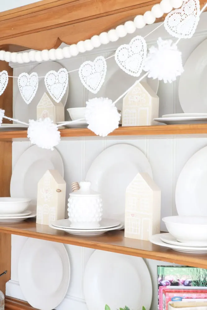 Decorate for Valentine with wooden gingerbread houses nestled on a hutch and garland of heart shaped doilies, felt ball garland and yarn ball garland all in white.