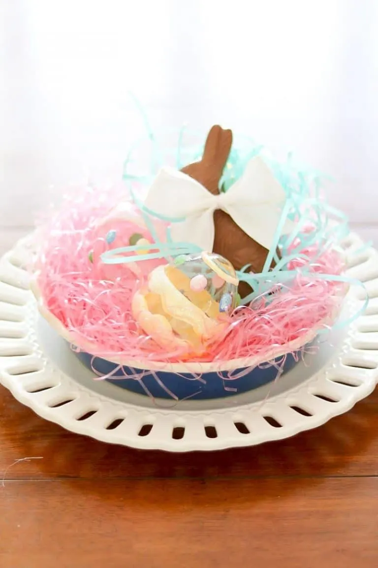 Easter centerpiece of layered dishes, bright colored grass, chocolate bunny, glass eggs.