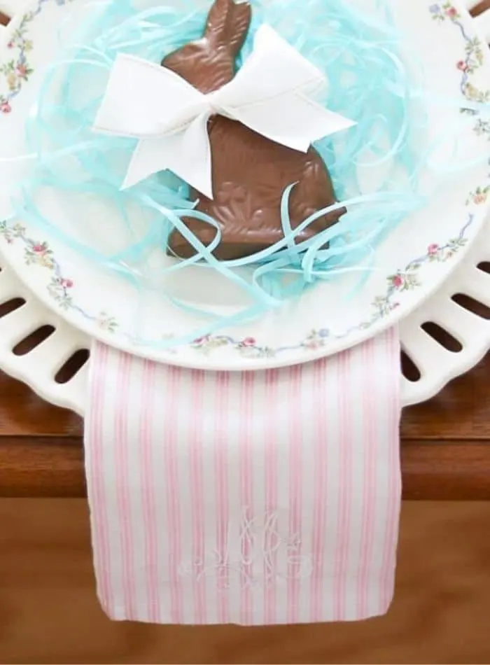 Easter place setting with l layered white dishes, delicate floral trimmed china, pink and white striped ticking dish towel monogrammed, blue Easter grass, chocolate bunny in the middle and a white bow.
