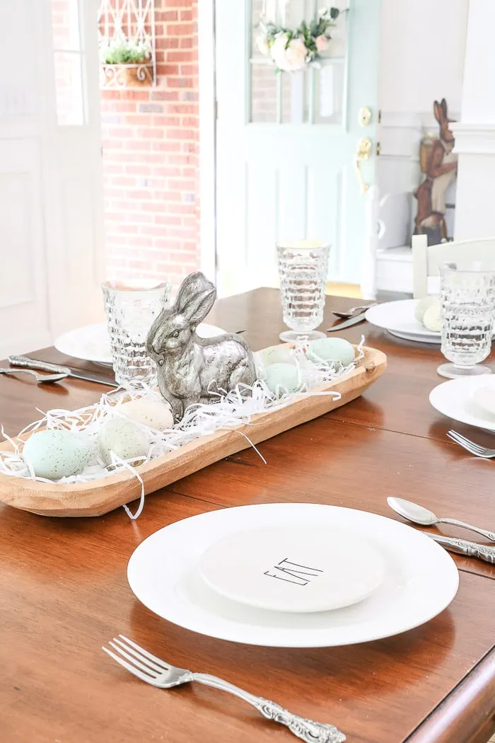 Easter centerpiece of a dough bowl filled with white grass, speckled eggs and a silver bunny mold in the middle.  And Rae Dunn Easter place settings.