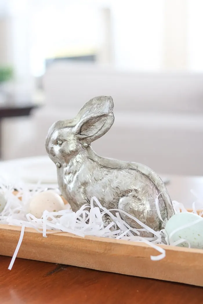 Easter centerpiece of a dough bowl filled with white grass, speckled eggs and a silver bunny mold in the middle.