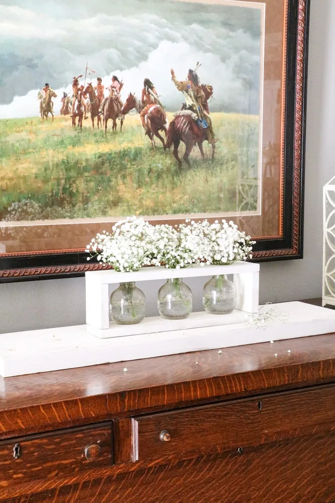 Baby's breath flowers arrangement into a wooden framed vase holder on a buffet in a dining room.