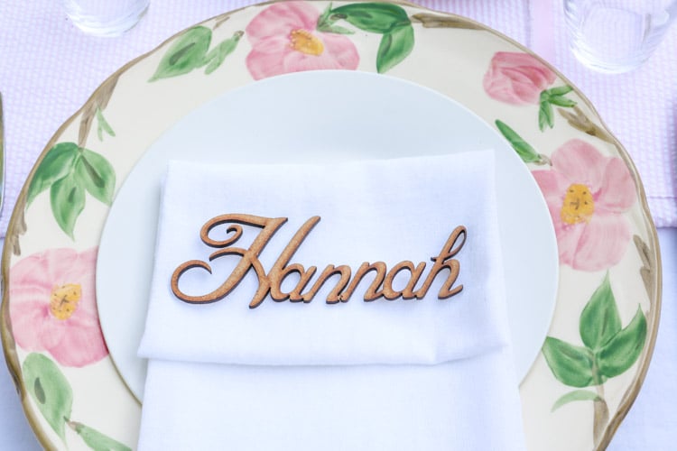 Garden party decoration ideas using white linen tablecloth, pink and white runner and desert rose dishes with a white salad plate in the middle with a folded linen napkin and a wooden laser cut name of guest.