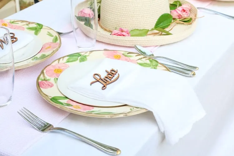 Garden party decoration ideas using white linen tablecloth, pink and white runner and desert rose dishes with a white salad plate in the middle with a folded linen napkin and a wooden laser cut name of guest.
