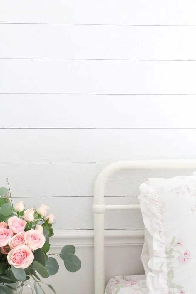 Shiplap peel and stick wallpaper on accent wall with a floral arrangement of flowers on a table sitting next to a white bed.
