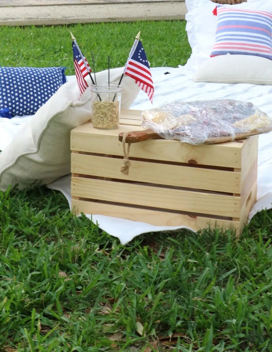 Fourth of July picnic ideas using a wooden crate turned upside down to act as a table.  Charcuterie board filled with food and a small mason jar filled with oatmeal that has two mini Betsy Ross flags inside.