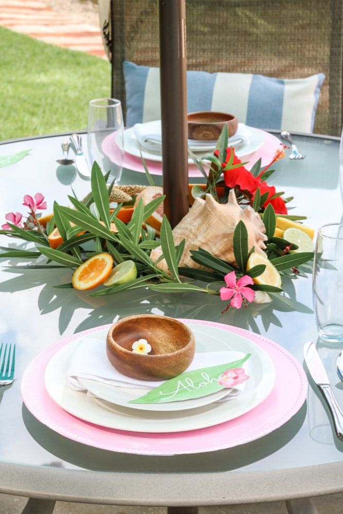 A Tiki party ideas with a bright center piece with shells, leafs, cut citrus, and colorful flowers. Along with arangged plates with a wooden bowl with Dole Whip.