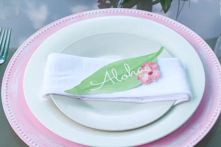 A Hawaiian table setting with a pink painted charger with a napkin wrapped salad plate with a leaf printable that says Aloha.
