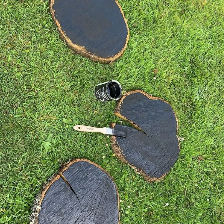 Slices of a wood log with raw edges are laying in the grass being painted with a black chalkboard paint.