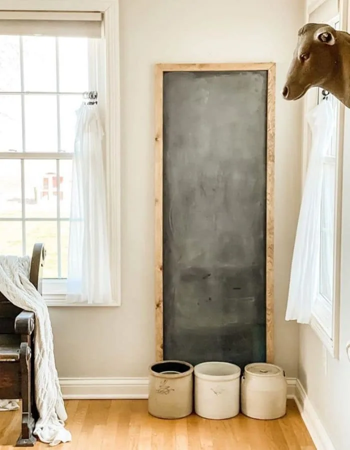 A vary tall farmhouse style chalk board leaning against a wall with 3 large crocks sitting at the base of the board.