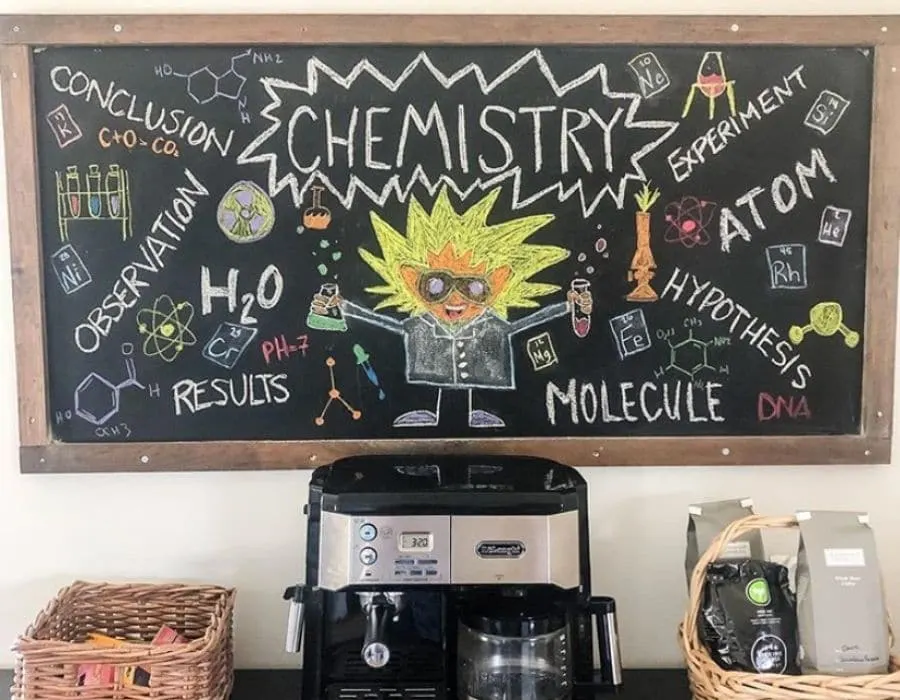 A long horizontal chalkboard for homeschool students, it is hanging on a wall above a coffee station with a coffee machine and some coffee as well.