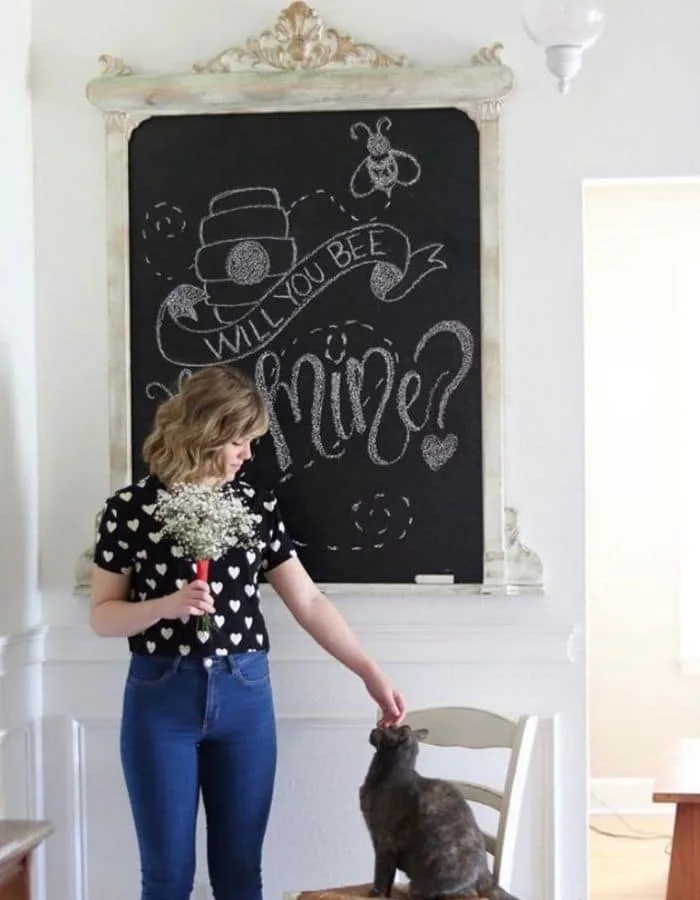 The chalkboard created in this post is behind a young woman holding a bouquet of baby breath flowers. She is gently petting her cat who is sitting in a chair next to her. The chalkboard says 'Will You Bee Mine?' written in chalk along with a drawing of a bee and its hive.