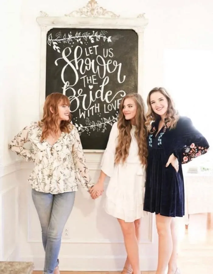 Three young woman holding hands in front of the chalkboard that was made in this post. The chalkboard says 'Let Us Shower The Bride With Love' written nicely in white chalk.