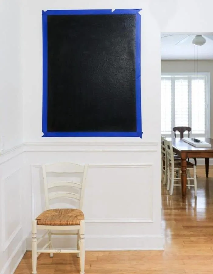 There is a small wall with a large rectangle of painters tape on the wall. Painted within the tape is a black chalkboard.