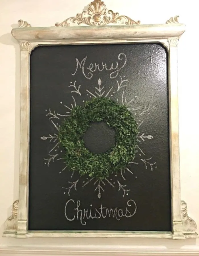 Homemade chalkboard that says 'Merry Christmas' on it written in cursive. In the center is a boxwood wreath with a pretty design boarding the wreath to resamble a snow flake.