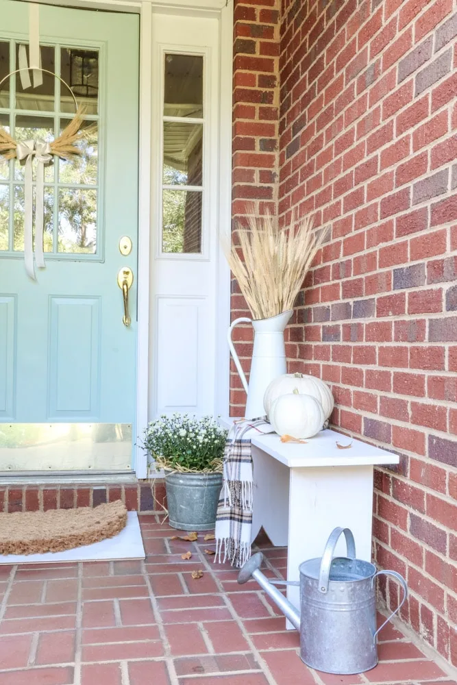 An autumn front door and porch with a little white bench, mums, pumpkins, and dried wheat.