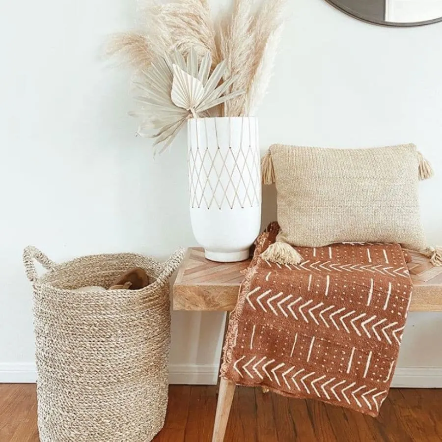 A wooden bench styled with a blanket, pillow, basket, and tall vase.