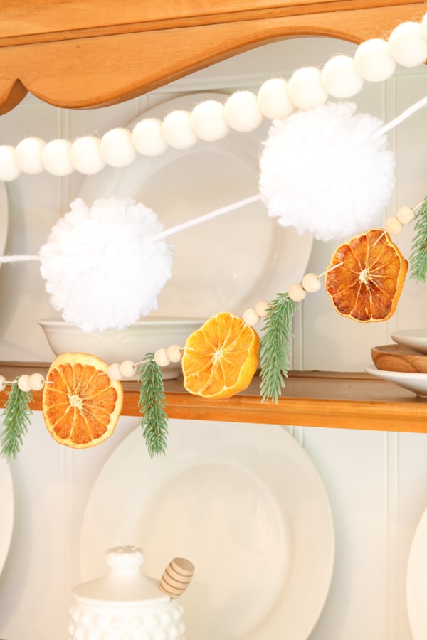 Dried orange garland with fir sprigs layered under a white felt ball and pom-pom garland on a hutch