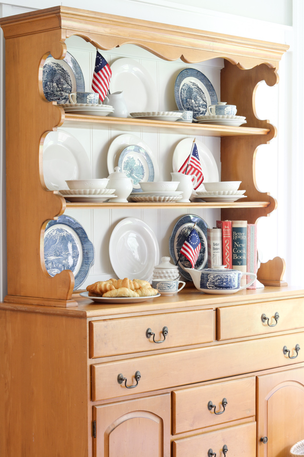 Decorating a hutch for 4th of July with Betsy Ross flags and blue Currier and Ives dishes