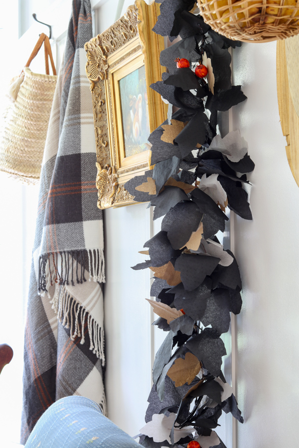 lack, white, and brown paper leaf garland hanging vertically on a wall of hooks with art, a neutral blanket, and a woven basket, cozy Halloween garland DIY idea