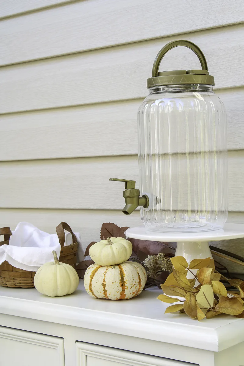 Drink dispenser sitting on cake stand with pumpkins and leaves as decoration