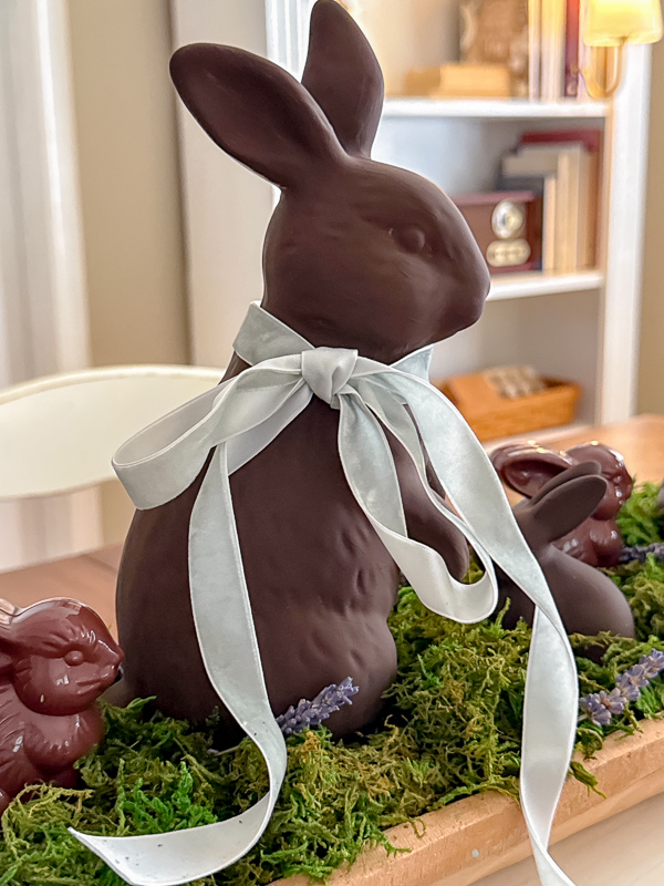 Ceramic bunny painted to look like a chocolate bunny with light blue ribbon around his neck and sitting in a bed of moss with other small brown bunnies around him.