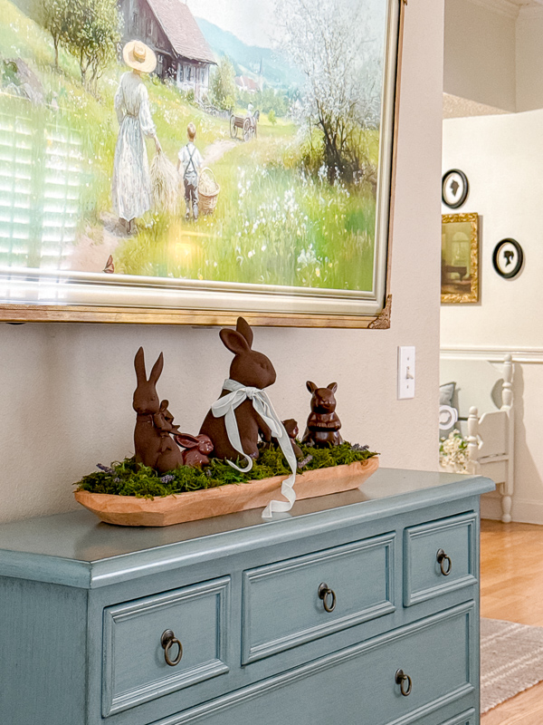 Faux chocolate bunny table decoration. Painted dark brown bunnies to look like chocolate rabbits sitting in a vintage dough bowl with moss and place on a table under a television