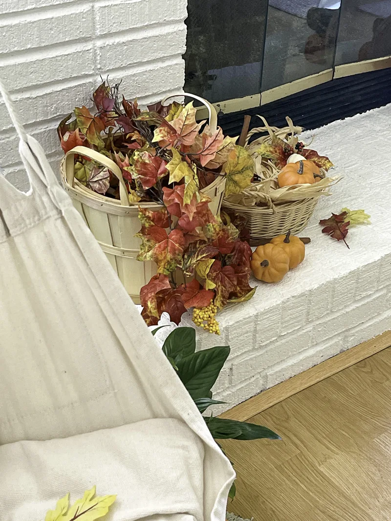 Decorating the hearth for fall with a bushel basket full of autumn leaves which is actually a garland and another basket filled with leaves and pumpkins.